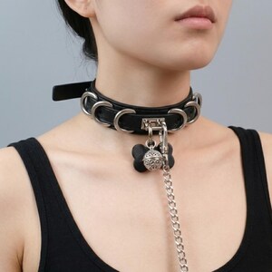 Puede incluir: Una gargantilla de cuero negro con múltiples anillos de metal plateado a lo largo de la banda. Un cierre central de metal plateado sostiene un dije negro en forma de hueso y una campana esférica de filigrana, con una cadena de metal plateado.