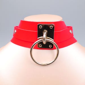 Puede incluir: Un collar choker rojo brillante con un gran anillo plateado y una placa metálica rectangular. El collar está hecho de un material liso y se asegura con remaches.