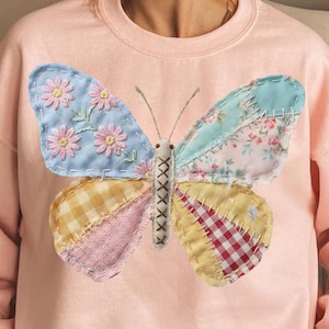 May include: A light pink sweatshirt featuring a large, colorful butterfly applique. The butterfly's wings are patchwork, with floral, gingham, and plaid patterns in shades of blue, pink, yellow, and red. The butterfly's body is stitched with a cross-stitch design.