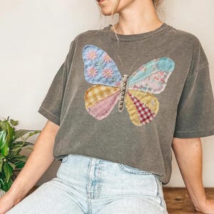May include: A gray t-shirt featuring a colorful butterfly applique. The butterfly wings are made of patchwork fabric in various patterns and colors, including blue with pink flowers, floral prints, and gingham. The shirt has a relaxed fit.