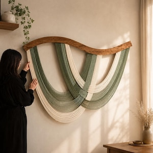 Op de afbeelding: Een wandhanger met een gebogen, natuurlijk gekleurde houten bovenbalk waaraan meerdere strengen saliegroen en crèmekleurig koord hangen. De koorden zijn gerangschikt in overlappende, U-vormige lussen die een gelaagd effect creëren.