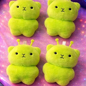 Pode incluir: Quatro brinquedos de pel&uacute;cia de urso alien&iacute;gena verde lim&atilde;o com olhos pretos e boca costurada. Cada brinquedo tem duas pequenas antenas e uma etiqueta branca. Os brinquedos est&atilde;o dispostos em uma superf&iacute;cie rosa e roxa brilhante.