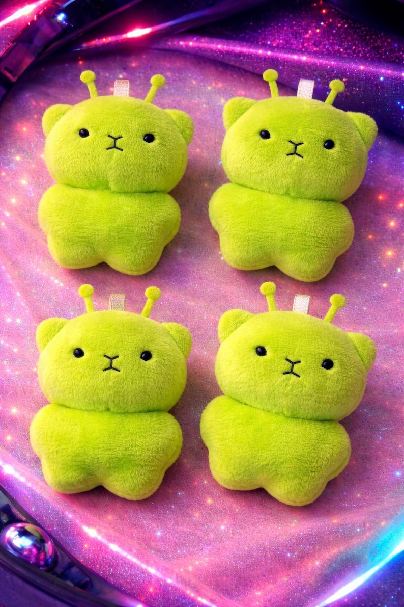 Pode incluir: Quatro brinquedos de pel&uacute;cia de urso alien&iacute;gena verde lim&atilde;o com olhos pretos e boca costurada. Cada brinquedo tem duas pequenas antenas e uma etiqueta branca. Os brinquedos est&atilde;o dispostos em uma superf&iacute;cie rosa e roxa brilhante.