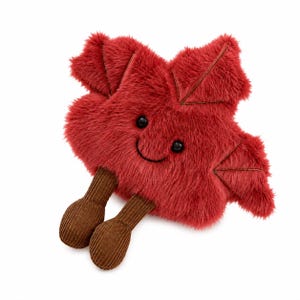 May include: A plush toy shaped like a red maple leaf with a soft, fuzzy texture. It features two small black bead eyes and a stitched smiling mouth. Two brown, corduroy-textured legs dangle from the bottom, ending in rounded, foot-like shapes.