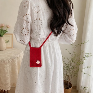 May include: A small, rectangular, red crocheted crossbody bag featuring a single pink and white flower appliqué on the front. The bag has a thin red strap and is worn over a white long-sleeved dress with a circular eyelet embroidery pattern.