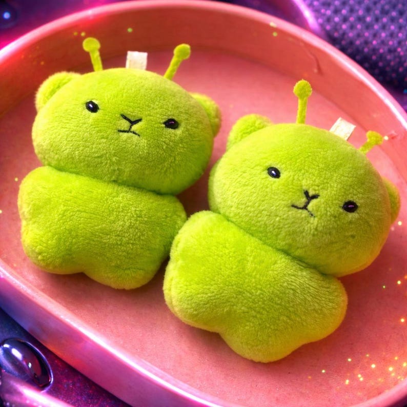 Pode incluir: Dois brinquedos de pel&uacute;cia verde lim&atilde;o, h&iacute;bridos de alien&iacute;gena e urso, com olhos pretos e boca costurada. Cada um tem duas antenas e uma etiqueta branca. Os brinquedos est&atilde;o em um recipiente rosa.