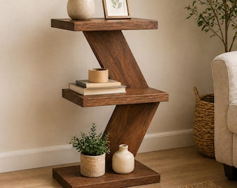 May include: A dark brown, zig-zag shaped wooden side table with three shelves. The top shelf holds a small potted plant and a framed picture. The middle shelf has books and a small container. The bottom shelf has a potted plant and a small vase.