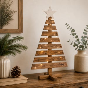 Op de afbeelding: Een decoratieve kerstboom gemaakt van horizontale donkerbruine houten latten in een driehoekige vorm, voorzien van een lichtgekleurde houten ster aan de top en dunne lichtsnoeren rondom de structuur, rustend op een houten kruisvoet.