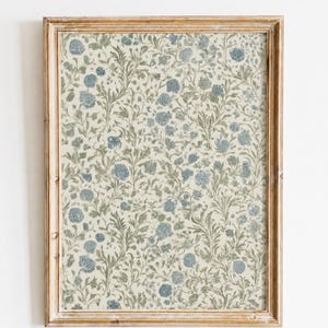 Puede incluir: Una impresión rectangular enmarcada con un patrón botánico repetitivo de flores azules y follaje verde salvia sobre un fondo crema. El marco está hecho de madera de color marrón claro con una veta visible que rodea la obra floral.
