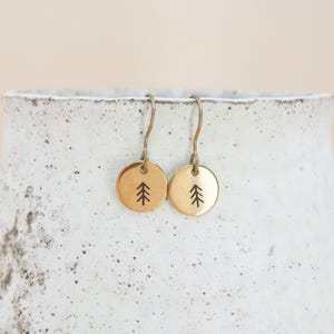 May include: A pair of gold-toned metal drop earrings featuring circular charms. Each charm is engraved with a simple black line drawing of a pine tree. The earrings are attached to curved metal ear wires for hanging.