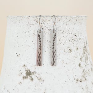 May include: A pair of silver-toned rectangular earrings. Each earring features a black floral design of a lavender sprig. The earrings hang from silver-colored hooks. The background is a textured white surface.