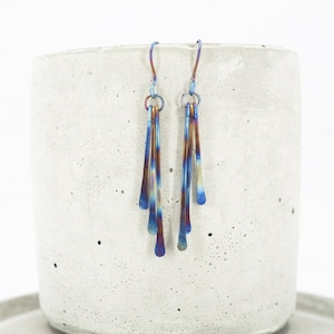 Puede incluir: Un par de pendientes colgantes con un degradado de color único. Los pendientes presentan una combinación de azul, bronce y morado. Los pendientes cuelgan de un pequeño aro y tienen forma de lágrima.