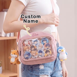 May include: A light pink crossbody bag with cat ears, a clear front display window filled with anime-style character buttons, and the name Eliza embroidered on the top. A plush fox charm hangs on the left and a plush penguin on the right.