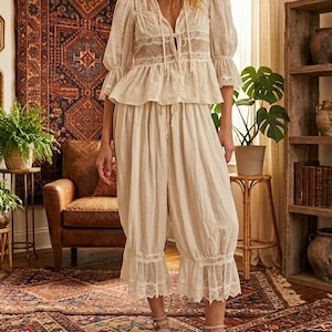 Op de afbeelding: Een vrouw draagt een crèmekleurige outfit in vintage-stijl. De set bestaat uit een blouse met kanten details en een taille met trekkoord, gecombineerd met een bijpassende cropped broek. De outfit is gecombineerd met beige sandalen.