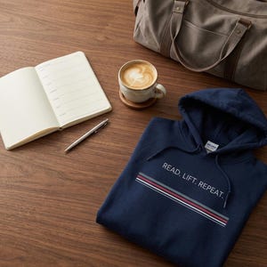 Peut inclure: Un sweat à capuche bleu marine avec le texte "READ. LIFT. REPEAT." en blanc. Le sweat à capuche a un design rayé rouge, blanc et gris. Un carnet, un stylo, une tasse de café, un dessous de verre et un sac de sport sont également sur la surface en bois.