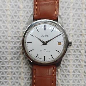 Peut inclure: Une montre-bracelet argentée vintage avec un cadran blanc et un bracelet en cuir marron. Le cadran affiche la marque "ENICAR", des aiguilles noires et le texte "17 STAR JEWELS". La montre a un affichage de la date à 3 heures.