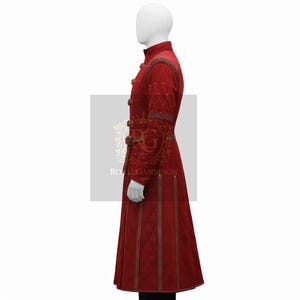 May include: A long red coat featuring a high collar, structured shoulders with braided trim, and a series of vertical brown leather-like straps extending down the front. The garment includes a waist belt and a quilted texture throughout.