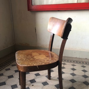 May include: A vintage wooden chair with a curved back and seat. The chair has a dark brown finish with visible wear and tear, suggesting age. The legs are sturdy and the overall design is simple and functional. The chair is placed on a patterned tile floor.