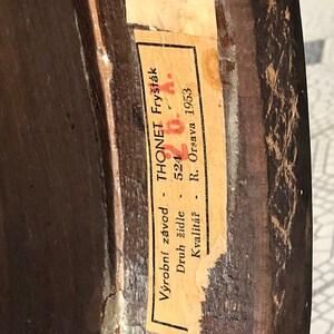 May include: Close-up of a vintage wooden furniture piece with a partially visible label. The label, in Czech, reads "THONET Fry&scaron;t&aacute;k" and includes the number "524" and the year "1953". The wood has a dark brown finish with visible grain and some wear.