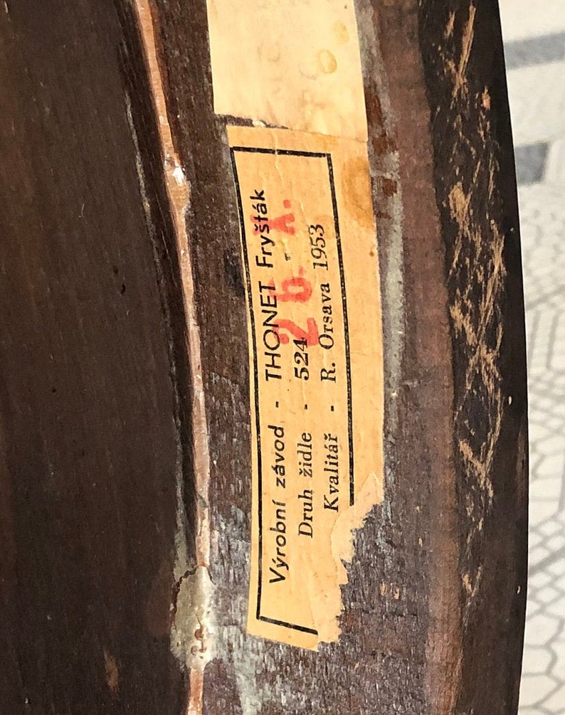 May include: Close-up of a vintage wooden furniture piece with a partially visible label. The label, in Czech, reads "THONET Fry&scaron;t&aacute;k" and includes the number "524" and the year "1953". The wood has a dark brown finish with visible grain and some wear.