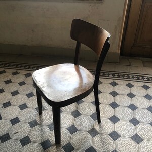 May include: A vintage wooden chair with a curved back and a dark-stained frame. The seat shows wear, with a light-colored finish. The chair stands on a patterned tile floor, with a window and door in the background. The chair is a classic piece of furniture.