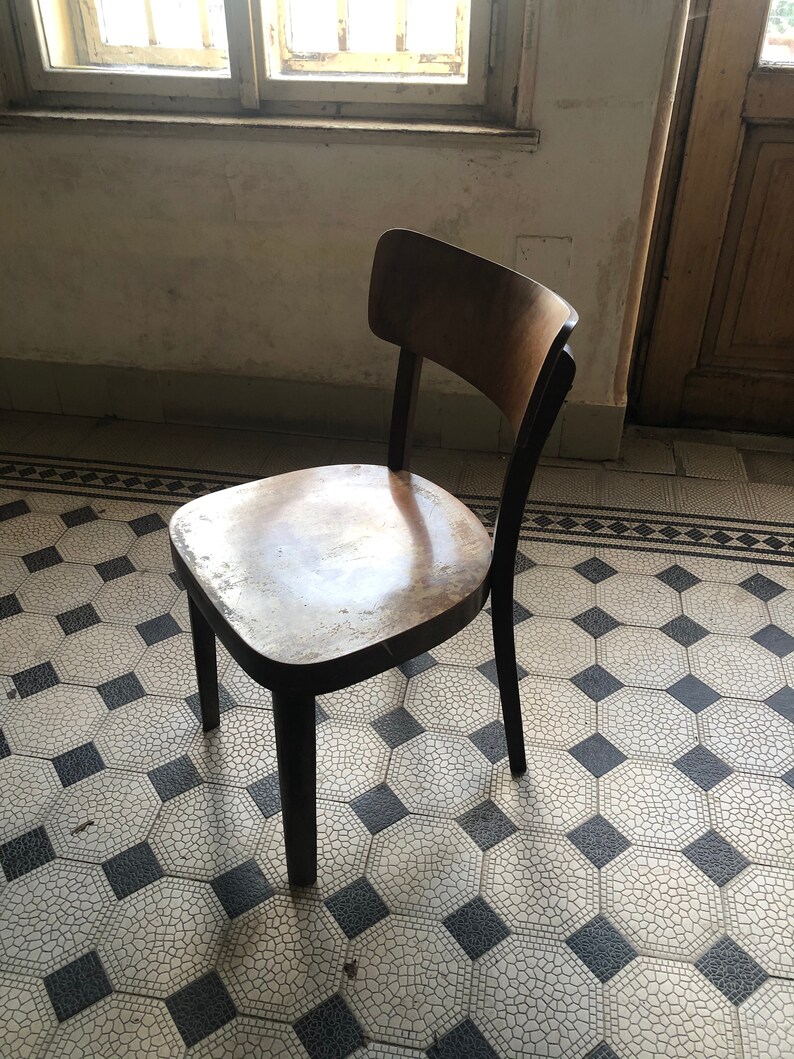 May include: A vintage wooden chair with a curved back and a dark-stained frame. The seat shows wear, with a light-colored finish. The chair stands on a patterned tile floor, with a window and door in the background. The chair is a classic piece of furniture.