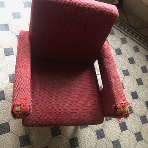 Op de afbeelding: Een versleten, rode fauteuil met beschadigde armleuningen. De stof heeft een geweven textuur. De stoel staat op een betegelde vloer met een patroon. De stoel lijkt vintage.
