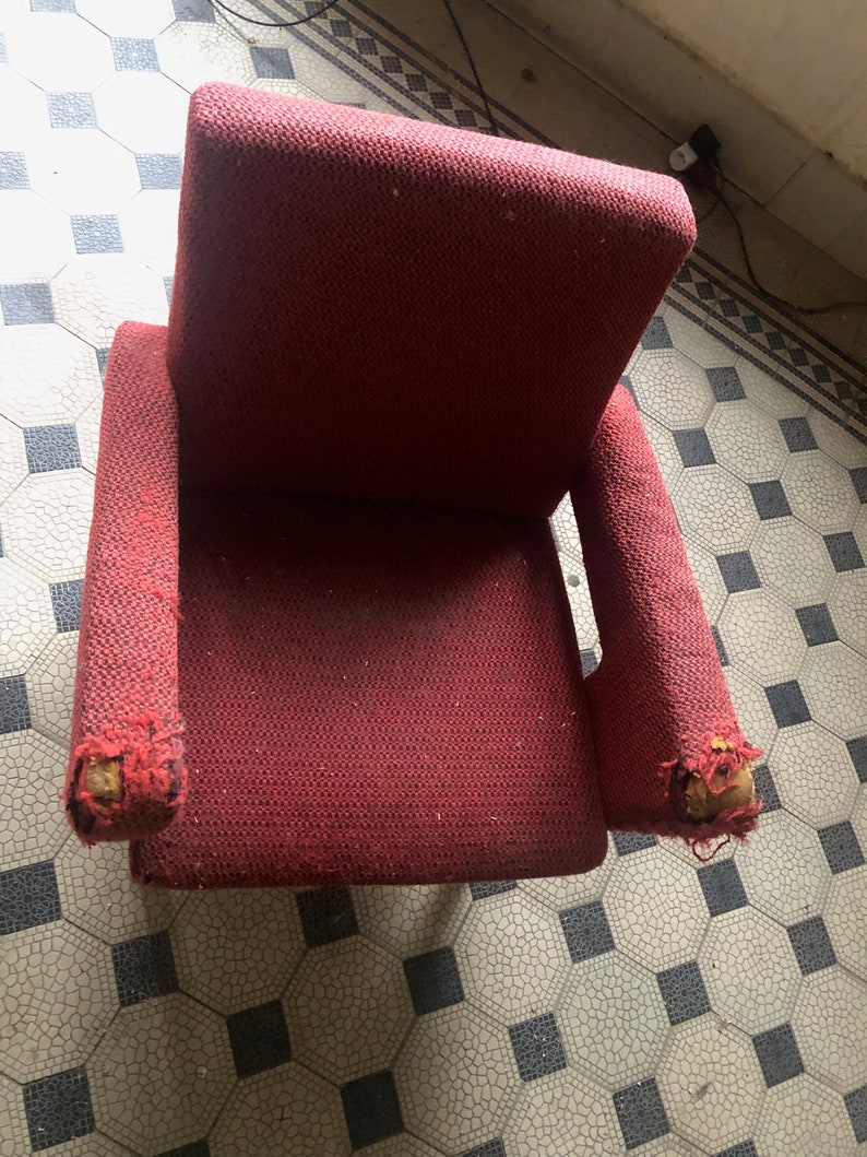 Op de afbeelding: Een versleten, rode fauteuil met beschadigde armleuningen. De stof heeft een geweven textuur. De stoel staat op een betegelde vloer met een patroon. De stoel lijkt vintage.