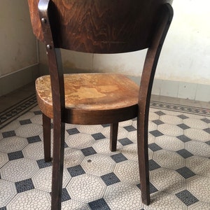 May include: A vintage wooden chair with a curved back and a worn seat. The chair is made of dark brown wood and has four legs. The back of the chair is attached to the seat with two metal rivets. The chair is on a patterned tile floor.