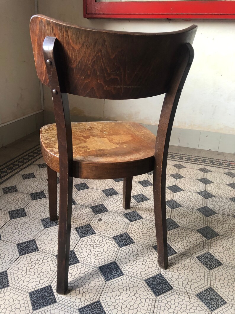 May include: A vintage wooden chair with a curved back and a worn seat. The chair is made of dark brown wood and has four legs. The back of the chair is attached to the seat with two metal rivets. The chair is on a patterned tile floor.