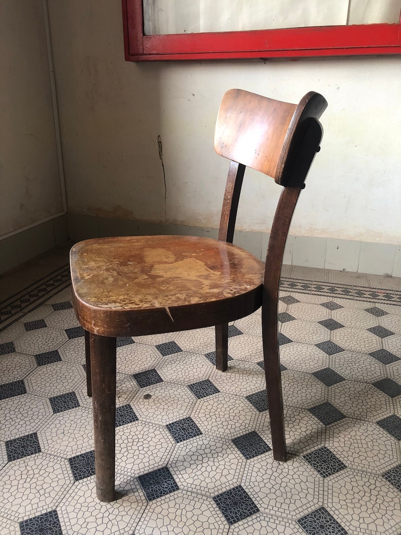 May include: A vintage wooden chair with a curved back and seat. The chair has a dark brown finish with visible wear and tear, suggesting age. The legs are sturdy and the overall design is simple and functional. The chair is placed on a patterned tile floor.