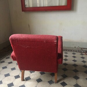 Op de afbeelding: Een vintage rode fauteuil met houten poten. De stoel heeft een textuurstof en staat tegen een witte muur met een rood omkaderd raam. De vloer heeft een patroon van tegels.