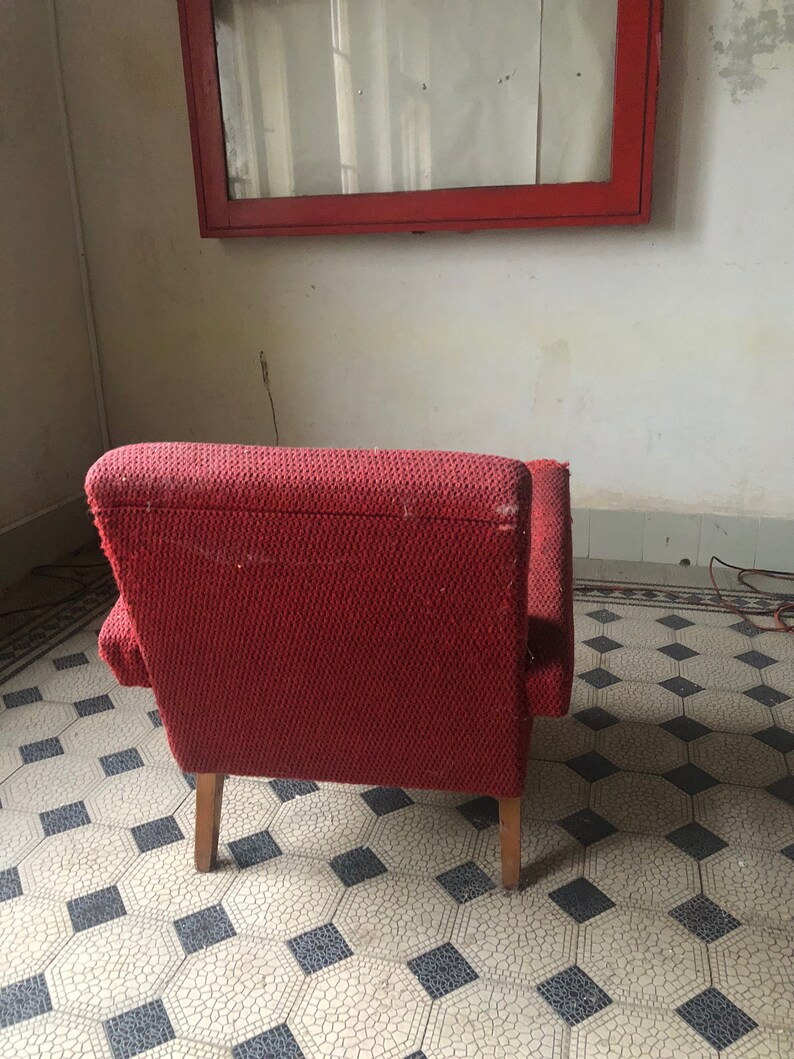 Op de afbeelding: Een vintage rode fauteuil met houten poten. De stoel heeft een textuurstof en staat tegen een witte muur met een rood omkaderd raam. De vloer heeft een patroon van tegels.