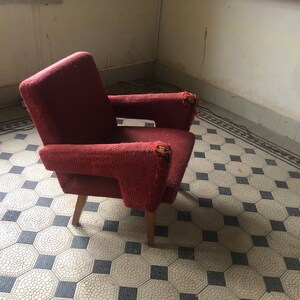 Op de afbeelding: Een vintage rode fauteuil met houten poten. De stoel heeft een textuurstof en vertoont slijtage aan de armleuningen. Het ontwerp is een klassieke mid-century stijl, geschikt voor een woonkamer of leeshoek.