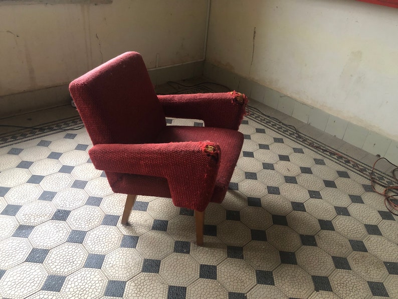 Op de afbeelding: Een vintage rode fauteuil met houten poten. De stoel heeft een textuurstof en vertoont slijtage aan de armleuningen. Het ontwerp is een klassieke mid-century stijl, geschikt voor een woonkamer of leeshoek.