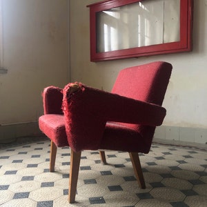Op de afbeelding: Een vintage rode fauteuil met houten poten. De stoel heeft een textuurstof en vertoont tekenen van slijtage op de armleuningen. Een rood omkaderde vitrine hangt aan de muur achter de stoel. De vloer heeft een geometrisch tegelpatroon.
