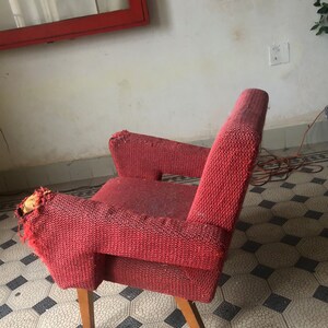 Op de afbeelding: Een vintage rode fauteuil met houten poten. De stoel heeft een textuurstof en vertoont tekenen van slijtage, met wat rafels op de armleuningen. Het ontwerp heeft een lage rugleuning en armleuningen, wat wijst op een mid-century modern stijl.