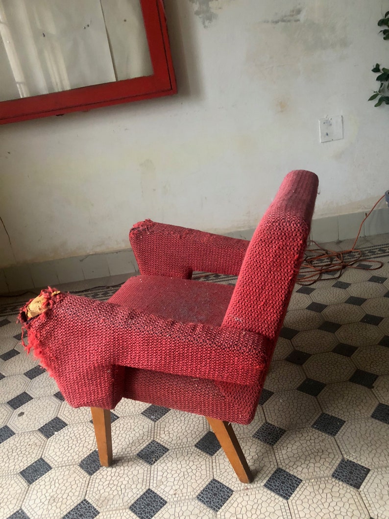 Op de afbeelding: Een vintage rode fauteuil met houten poten. De stoel heeft een textuurstof en vertoont tekenen van slijtage, met wat rafels op de armleuningen. Het ontwerp heeft een lage rugleuning en armleuningen, wat wijst op een mid-century modern stijl.