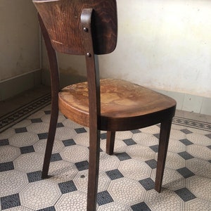 May include: A vintage wooden chair with a curved back and seat. The chair is made of dark-stained wood and shows signs of wear. The backrest is attached with metal rivets. The chair sits on a patterned tile floor.