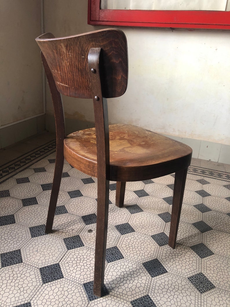 May include: A vintage wooden chair with a curved back and seat. The chair is made of dark-stained wood and shows signs of wear. The backrest is attached with metal rivets. The chair sits on a patterned tile floor.