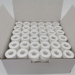 May include: A white cardboard box filled with numerous white spools of thread. The spools are neatly arranged, showcasing a consistent cylindrical shape with a central hole. The box is open, revealing the contents.