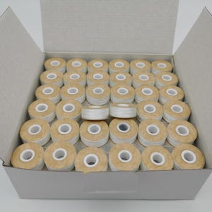 May include: A cardboard box filled with numerous spools of white thread. The spools have a tan-colored exterior and a white center. The box is open, revealing the neatly arranged spools.