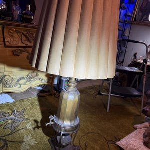 Puede incluir: Una lámpara de mesa con una pantalla de tela color canela, alta y plisada, sobre una base metálica de tono latón con una manija de grifo decorativa. La lámpara se apoya en una base de madera oscura de tres patas e incluye un cable eléctrico marrón.