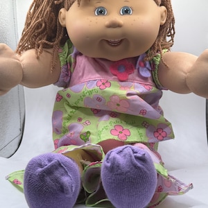 May include: A Cabbage Patch Kids doll with brown yarn hair, blue eyes, and freckles. The doll wears a light green and pink floral patterned dress with a small purple flower accent on the chest and solid purple socks on its feet.