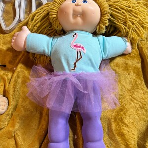 Puede incluir: Una muñeca con cabello de hilo dorado, ojos azules y un diente visible. La muñeca viste una camiseta de manga larga azul claro con un gráfico de flamenco rosa, un tutú de tul morado y pantalones de tela morados a juego que cubren las piernas.