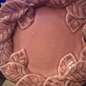 Puede incluir: Un plato redondo de cerámica en color rosa empolvado con un borde en relieve de hojas superpuestas. El centro del plato tiene una superficie lisa y esmaltada con un acabado craquelado sutil en toda la pieza.