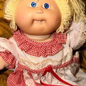 Op de afbeelding: Een pop met een lichte huid, helderblauwe ogen en blond garen haar. De pop heeft één zichtbare tand en draagt een witte jurk met rood-wit geblokte randen, een rucheskraag, broderie anglaise details en een dun rood lint in de taille.
