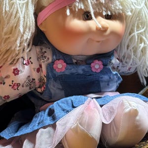 Pode incluir: Uma boneca com cabelo loiro de lã, uma faixa rosa com um laço e bochechas rosadas. A boneca veste uma camisa branca de manga comprida com um pequeno padrão floral e um vestido jardineira de ganga azul com dois botões rosa em forma de flor.