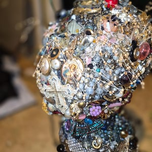 Puede incluir: Un objeto decorativo cubierto por un denso mosaico de joyas vintage variadas, incluyendo pedrería transparente, cuentas de colores, botones y un colgante religioso central con un retrato y una cruz de tono plateado.