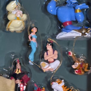 Puede incluir: Juego de adornos navideños con temática de Aladdin. Incluye figuras de Genio, Jasmine, Aladdin, Jafar, Abu, Iago y Rajah. Cada adorno es de colores brillantes y detallado, con cuerdas doradas para colgar. Para fans de Disney.